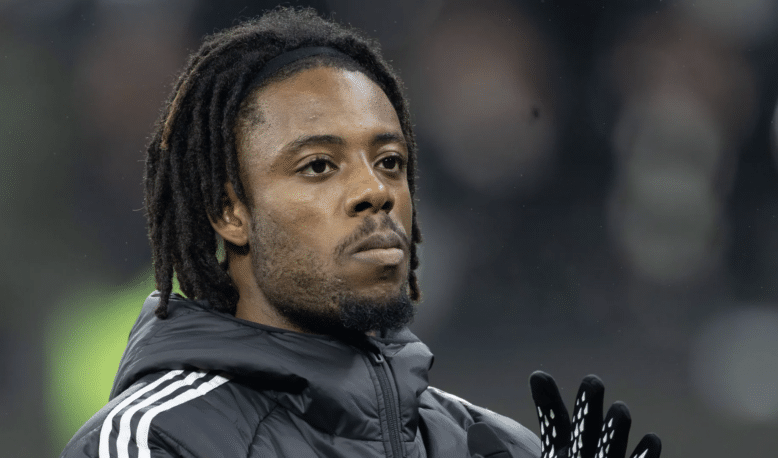 Close-up of a Black man with dreadlocks wearing a dark jacket and gloves, looking upward.