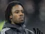 Close-up of a Black man with dreadlocks wearing a dark jacket and gloves, looking upward.
