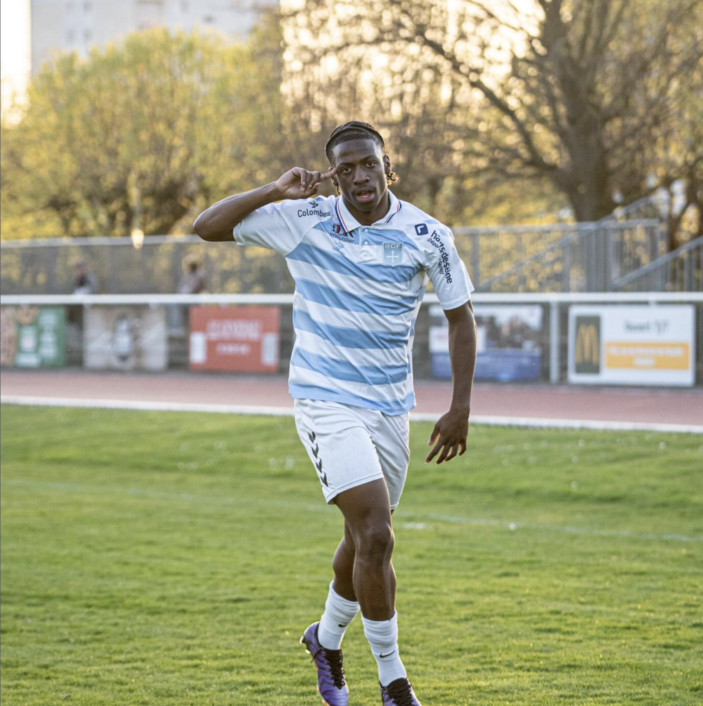 Capture décran 2026-07-11 à 214500 | AllezPaillade.com Soccer player in a light blue and white striped jersey running on a green field with trees and advertising boards in the background.