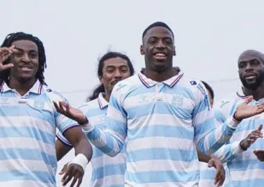 Five male athletes in light blue and white striped jerseys pose together and smile on a sports field, signaling victory.