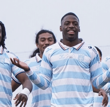 Five male athletes in light blue and white striped jerseys pose together and smile on a sports field, signaling victory.