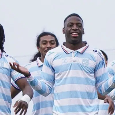 Five male athletes in light blue and white striped jerseys pose together and smile on a sports field, signaling victory.
