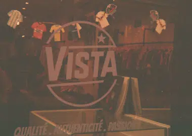 Interior of a clothing store with a large VISTA logo and circular emblem, shirts on racks in the background and a front display shelf in the foreground.