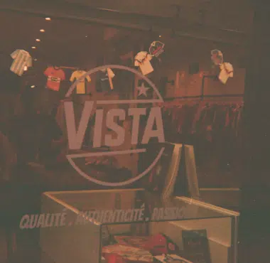 Interior of a clothing store with a large VISTA logo and circular emblem, shirts on racks in the background and a front display shelf in the foreground.