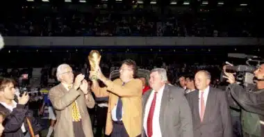 FRAC34172_COL671_01 | AllezPaillade.com Group of suited men raising a trophy on a sports field while photographers film and a cheering crowd watches in the stands.