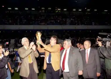 FRAC34172_COL671_01 | AllezPaillade.com Group of suited men raising a trophy on a sports field while photographers film and a cheering crowd watches in the stands.