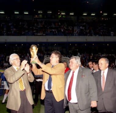 Group of suited men raising a trophy on a sports field while photographers film and a cheering crowd watches in the stands.