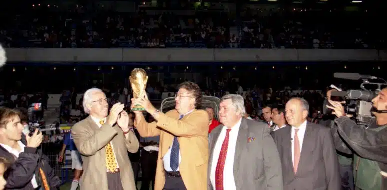 FRAC34172_COL671_01 | AllezPaillade.com Group of suited men raising a trophy on a sports field while photographers film and a cheering crowd watches in the stands.