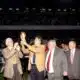 Group of suited men raising a trophy on a sports field while photographers film and a cheering crowd watches in the stands.