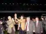 Group of suited men raising a trophy on a sports field while photographers film and a cheering crowd watches in the stands.