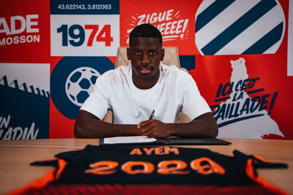 Grégory Ayem signature 2 | AllezPaillade.com Man in a white t-shirt sits at a desk, signing a document, against a bright red and blue graphic backdrop.