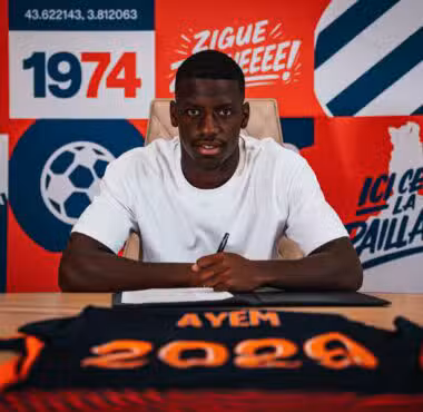 Man in a white t-shirt sits at a desk, signing a document, against a bright red and blue graphic backdrop.