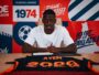 Man in a white t-shirt sits at a desk, signing a document, against a bright red and blue graphic backdrop.