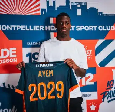 Young man wearing a white shirt holds up a dark blue sports jersey with orange '2029' and the name AYEM.