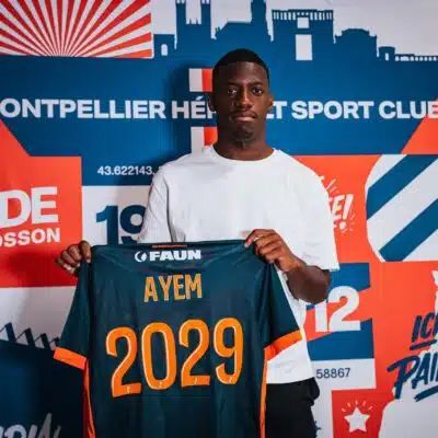 Young man wearing a white shirt holds up a dark blue sports jersey with orange '2029' and the name AYEM.
