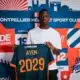 Young man wearing a white shirt holds up a dark blue sports jersey with orange '2029' and the name AYEM.