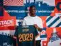 Young man wearing a white shirt holds up a dark blue sports jersey with orange '2029' and the name AYEM.