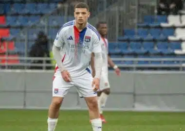 HDehauUXgAEumHJ-e1774000792937-770x433 | AllezPaillade.com Soccer player in a Lyon jersey standing on a green field with a stadium background, ready to play.