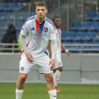 Soccer player in a Lyon jersey standing on a green field with a stadium background, ready to play.
