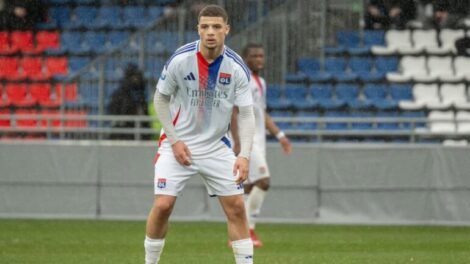 Soccer player in a Lyon jersey standing on a green field with a stadium background, ready to play.