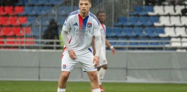 HDehauUXgAEumHJ-e1774000792937-770x433 | AllezPaillade.com Soccer player in a Lyon jersey standing on a green field with a stadium background, ready to play.