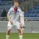 Soccer player in a Lyon jersey standing on a green field with a stadium background, ready to play.