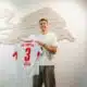 Man in a white T-shirt smiles while holding a white RB Leipzig jersey with the number 3, in front of a pale wall sculpture.