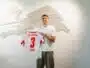 Man in a white T-shirt smiles while holding a white RB Leipzig jersey with the number 3, in front of a pale wall sculpture.