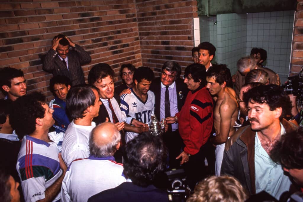 Montpellier HSC v Racing Paris - French Cup Final 1990 | AllezPaillade.com Crowd of men in sports jerseys surrounding a silver trophy during a celebration in a brick-walled room.