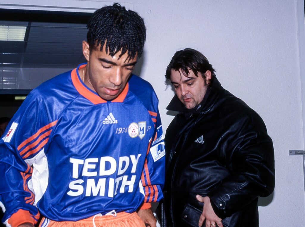 Patrice Loko Laurent Nicollin | AllezPaillade.com Two men indoors, one in a blue sports jersey with orange accents and large 'TEDDY SMITH' text, the other in a black jacket, both looking downward.