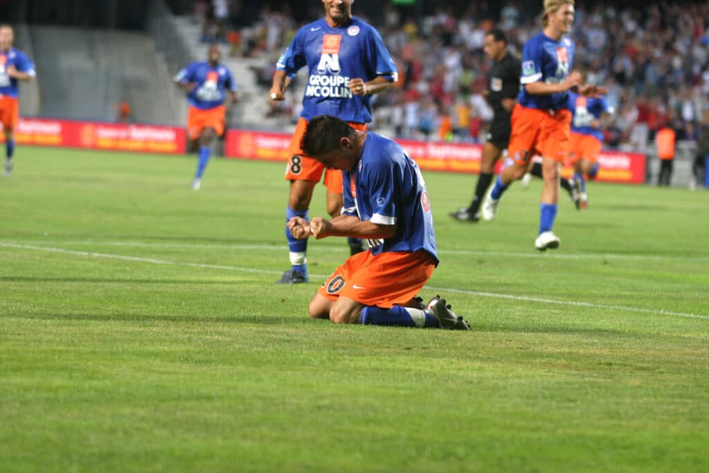 Fatih ATIK | AllezPaillade.com Soccer player in blue jersey kneels on grass as teammates run in the background. Move conveys a moment of celebration or injury after a play.