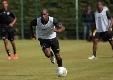 Soccer player in a white training vest dribbles the ball on a grassy field, teammates blurred in the background during practice.]','Soccer practice scene with a central player in a white vest chasing the ball on a sunny day.