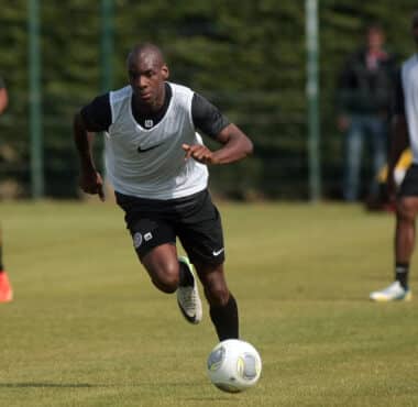 Soccer player in a white training vest dribbles the ball on a grassy field, teammates blurred in the background during practice.]','Soccer practice scene with a central player in a white vest chasing the ball on a sunny day.