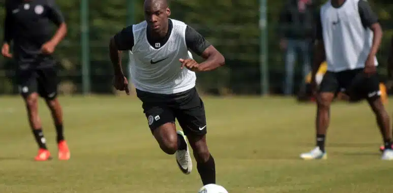 Soccer player in a white training vest dribbles the ball on a grassy field, teammates blurred in the background during practice.]','Soccer practice scene with a central player in a white vest chasing the ball on a sunny day.