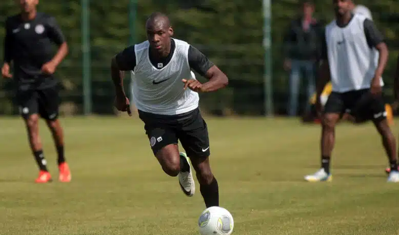 Soccer player in a white training vest dribbles the ball on a grassy field, teammates blurred in the background during practice.]','Soccer practice scene with a central player in a white vest chasing the ball on a sunny day.