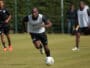 Soccer player in a white training vest dribbles the ball on a grassy field, teammates blurred in the background during practice.]','Soccer practice scene with a central player in a white vest chasing the ball on a sunny day.