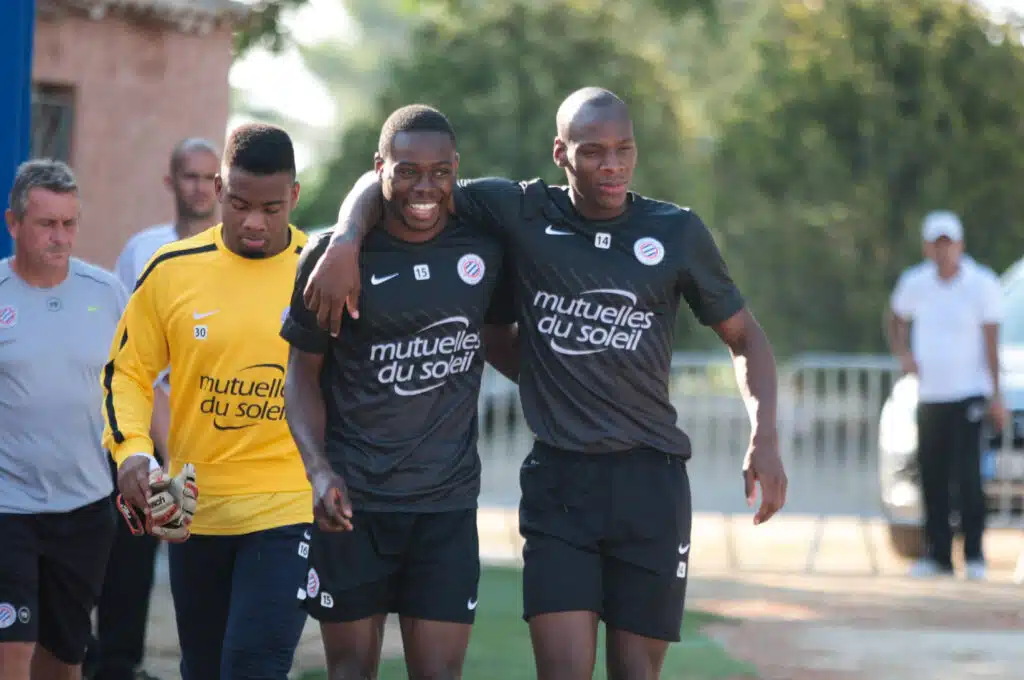 Group of soccer players with arms around each other, wearing dark jerseys with a sponsor logo, smiling outdoors.