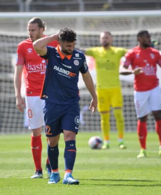 Soccer player in blue with a hand on his head, other players in red jerseys nearby, and a yellow-clad goalkeeper near the goal behind him.