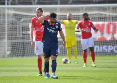 Soccer player in blue with a hand on his head, other players in red jerseys nearby, and a yellow-clad goalkeeper near the goal behind him.