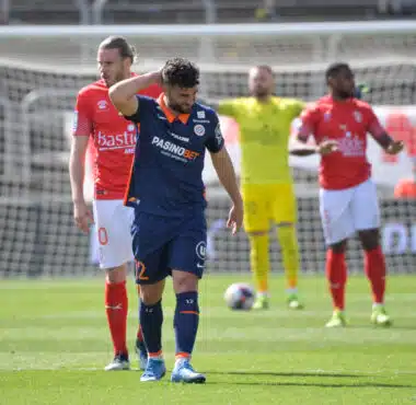 Soccer player in blue with a hand on his head, other players in red jerseys nearby, and a yellow-clad goalkeeper near the goal behind him.