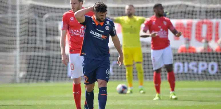Jordan Ferri Nimes Olympique v Montpellier HSC - Ligue 1 Uber Eats | AllezPaillade.com Soccer player in blue with a hand on his head, other players in red jerseys nearby, and a yellow-clad goalkeeper near the goal behind him.