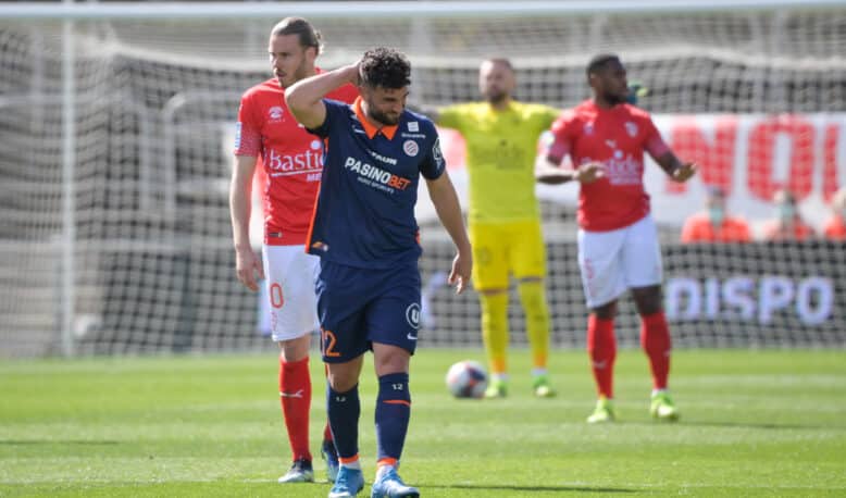 Soccer player in blue with a hand on his head, other players in red jerseys nearby, and a yellow-clad goalkeeper near the goal behind him.
