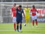Soccer player in blue with a hand on his head, other players in red jerseys nearby, and a yellow-clad goalkeeper near the goal behind him.