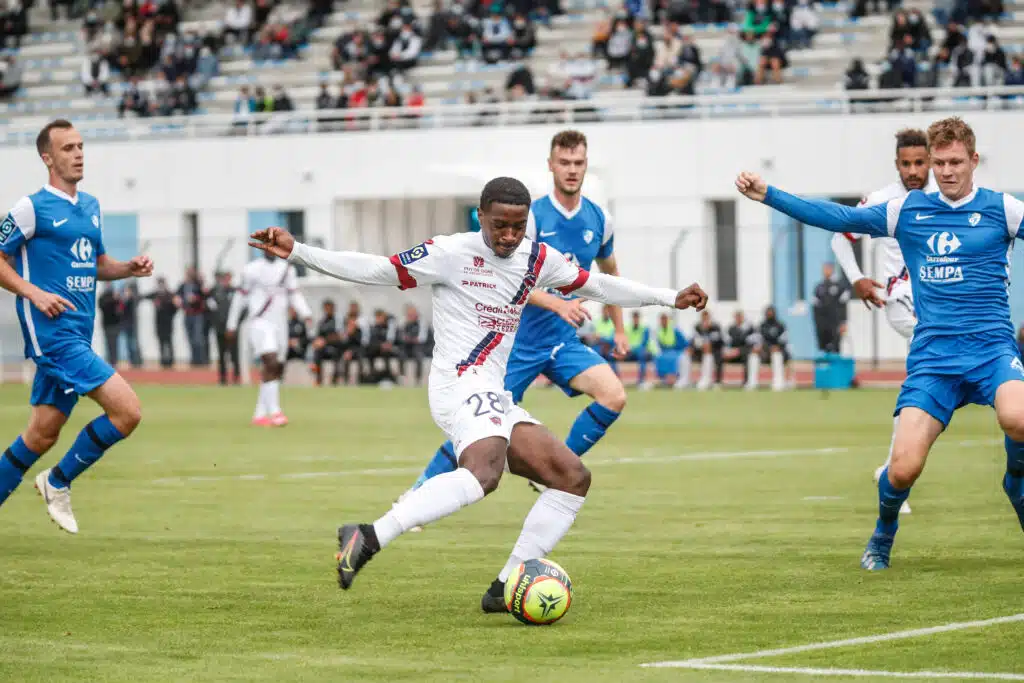 Grenoble v Clermont - Friendly match | AllezPaillade.com Soccer player in white with the ball dribbles past blue-clad opponents on a turf field with a stadium crowd in the background