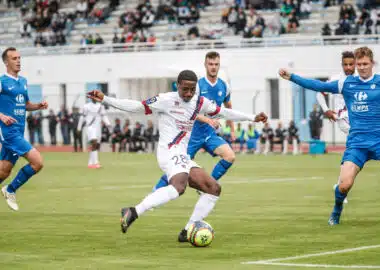 Grenoble v Clermont - Friendly match | AllezPaillade.com Soccer player in white with the ball dribbles past blue-clad opponents on a turf field with a stadium crowd in the background