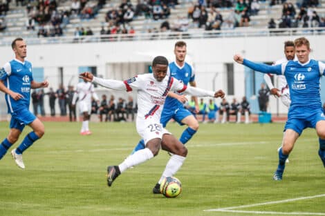 Soccer player in white with the ball dribbles past blue-clad opponents on a turf field with a stadium crowd in the background