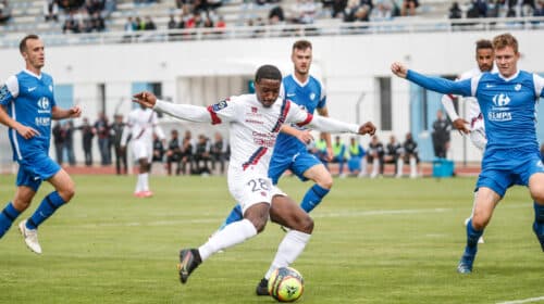 Soccer player in white with the ball dribbles past blue-clad opponents on a turf field with a stadium crowd in the background