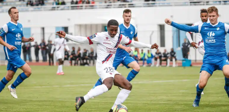 Grenoble v Clermont - Friendly match | AllezPaillade.com Soccer player in white with the ball dribbles past blue-clad opponents on a turf field with a stadium crowd in the background