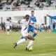 Soccer player in white with the ball dribbles past blue-clad opponents on a turf field with a stadium crowd in the background