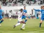 Soccer player in white with the ball dribbles past blue-clad opponents on a turf field with a stadium crowd in the background